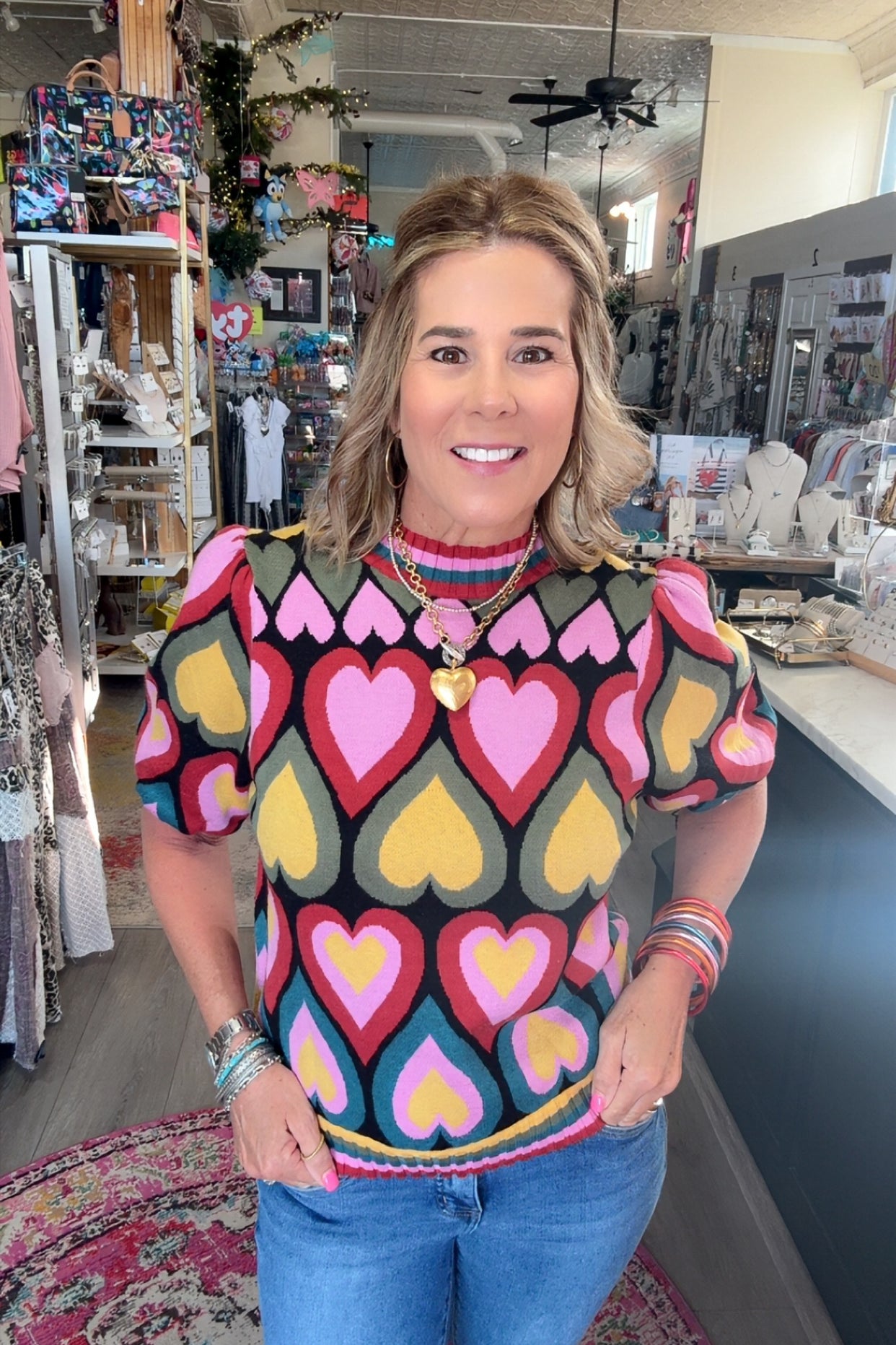 A woman wearing a colorful short-sleeve knit top with a heart pattern, standing in a retail store.