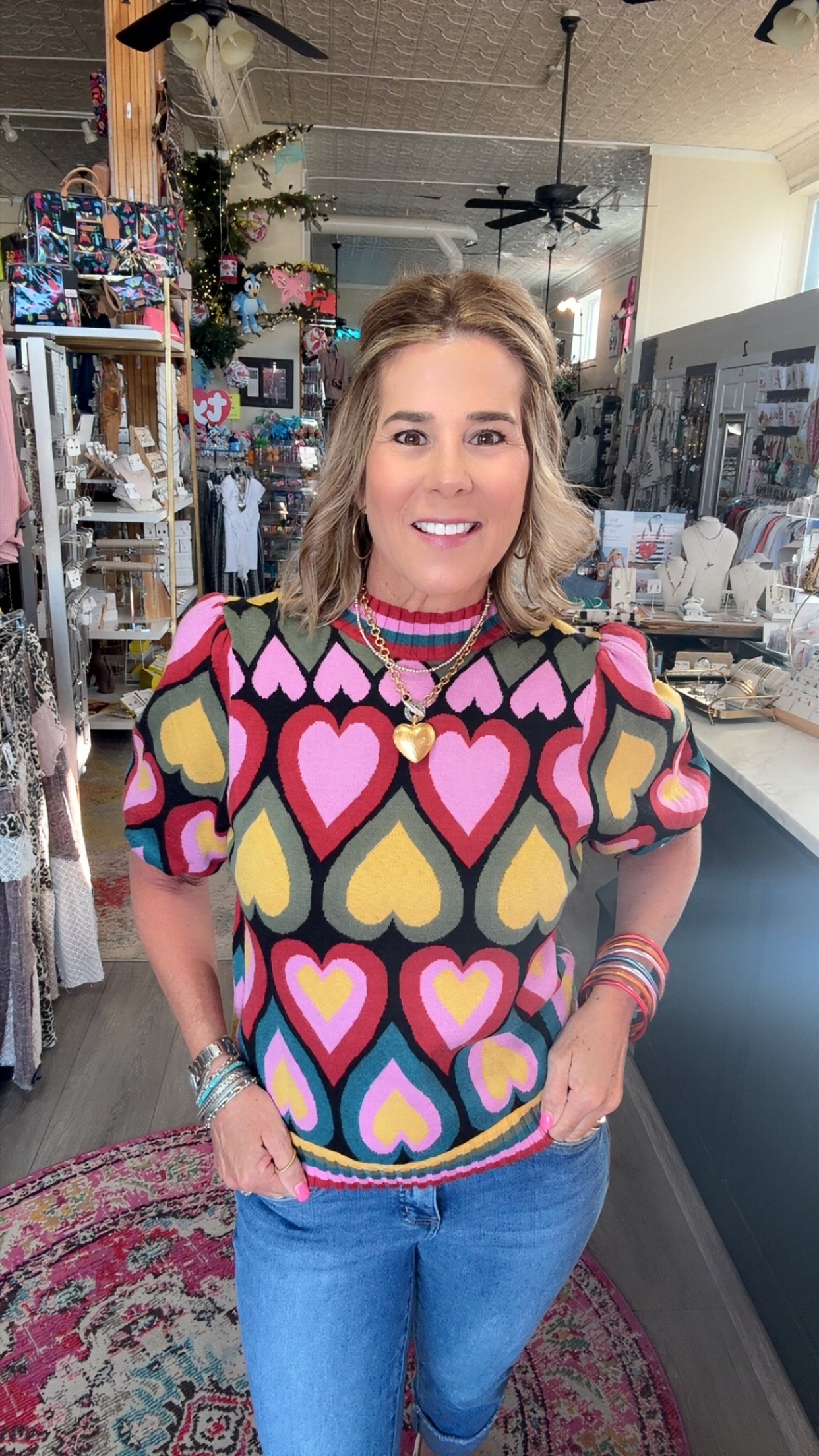 A woman wearing a colorful short-sleeve knit top with a heart pattern, standing in a retail store.