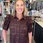A woman wearing a dark brown western-style puff-sleeve shirt with contrast stitching and blue jeans, standing inside a retail store.