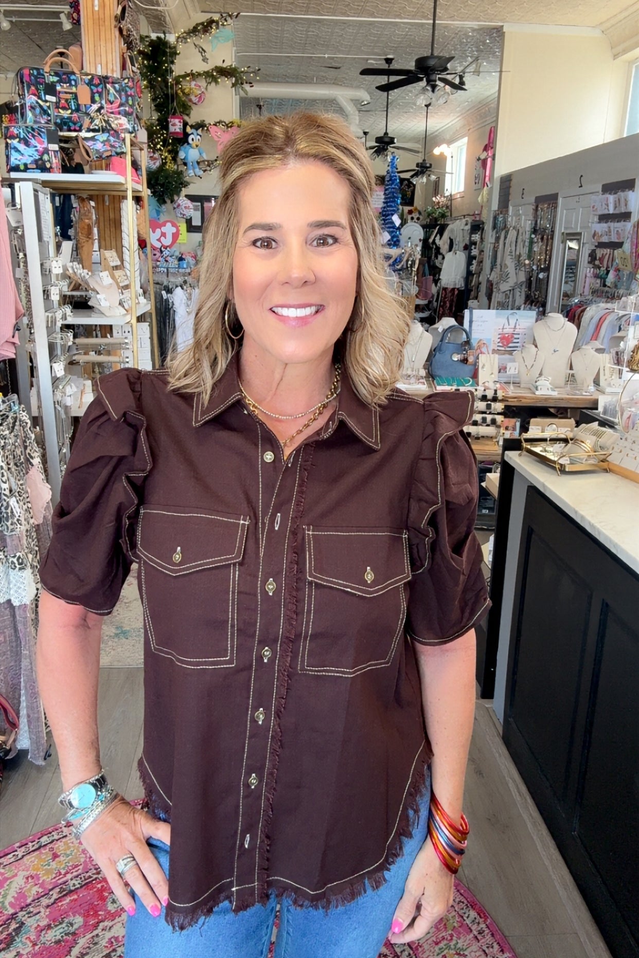 A woman wearing a dark brown western-style puff-sleeve shirt with contrast stitching and blue jeans, standing inside a retail store.