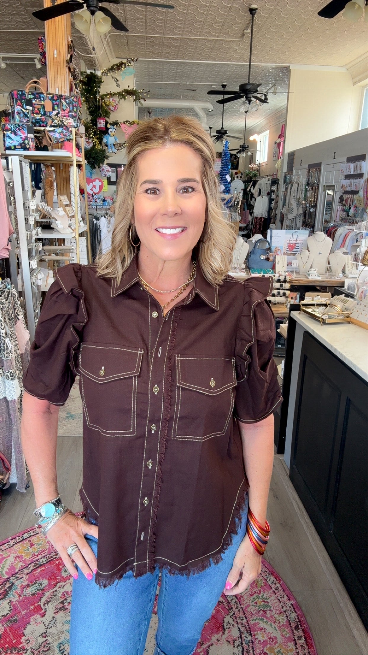 A woman wearing a dark brown western-style puff-sleeve shirt with contrast stitching and blue jeans, standing inside a retail store.