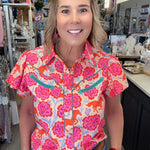 A woman wearing a vibrant short-sleeve western-style button-up shirt with a pink, orange, and blue floral pattern, paired with blue jeans and a brown leather belt, standing inside a retail store.