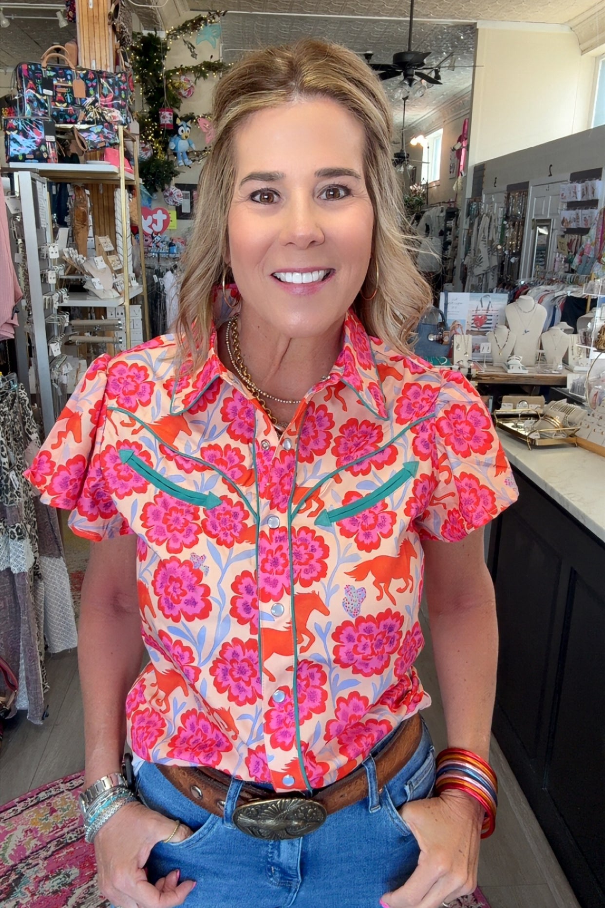 A woman wearing a vibrant short-sleeve western-style button-up shirt with a pink, orange, and blue floral pattern, paired with blue jeans and a brown leather belt, standing inside a retail store.