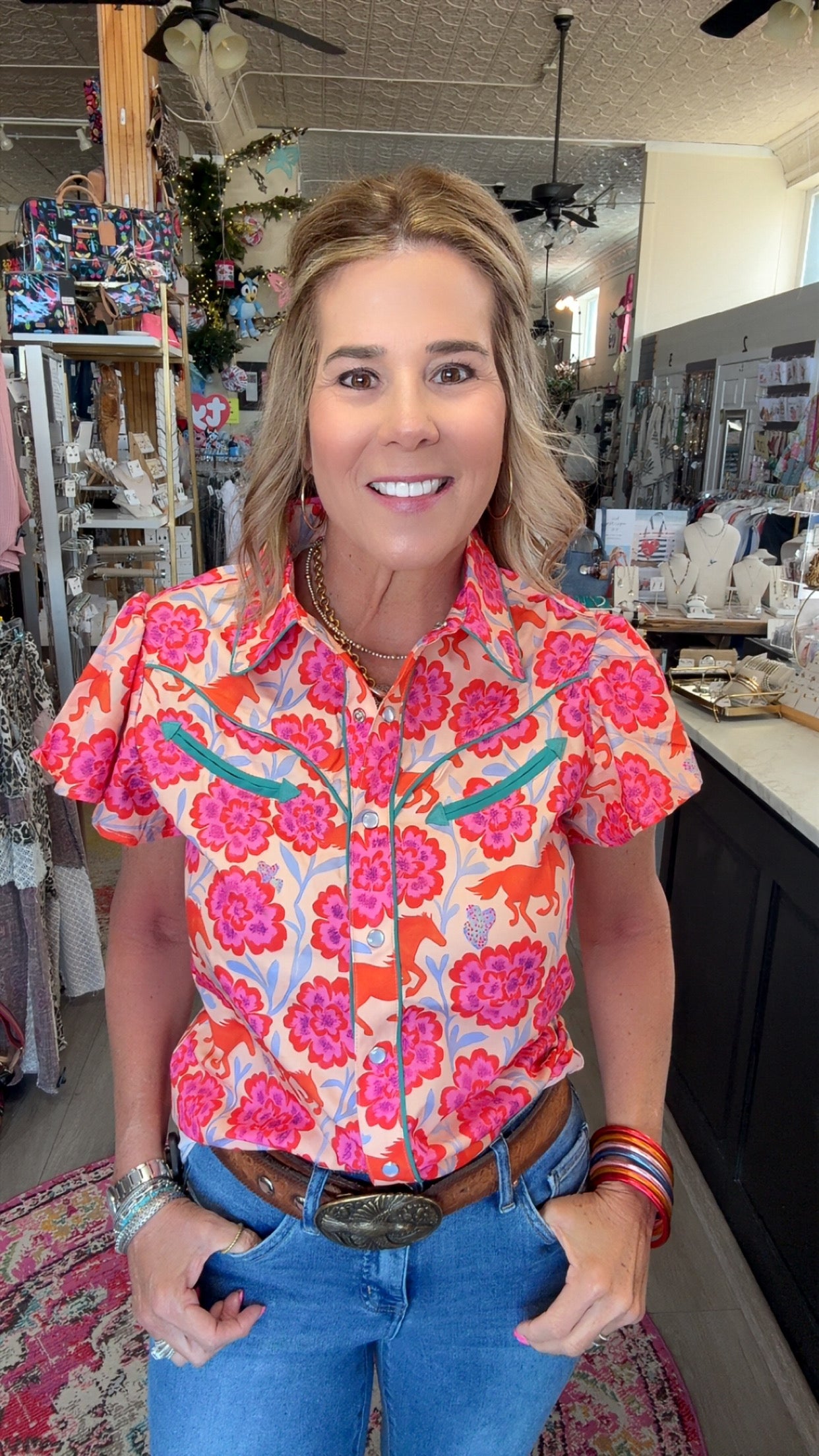 A woman wearing a vibrant short-sleeve western-style button-up shirt with a pink, orange, and blue floral pattern, paired with blue jeans and a brown leather belt, standing inside a retail store.