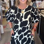 A woman modeling a short puff-sleeve dress with a bold black and white organic spot pattern, standing inside a boutique shop.