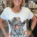 A woman smiling inside a retail shop, wearing a white graphic t-shirt with a detailed illustration of a tiger wearing a cowboy hat, paired with blue jeans.