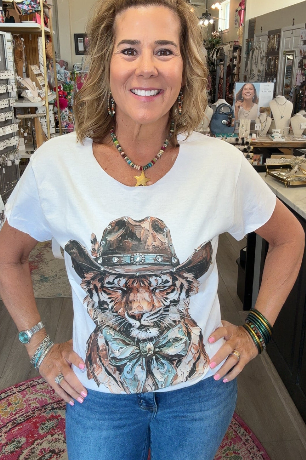 A woman smiling inside a retail shop, wearing a white graphic t-shirt with a detailed illustration of a tiger wearing a cowboy hat, paired with blue jeans.