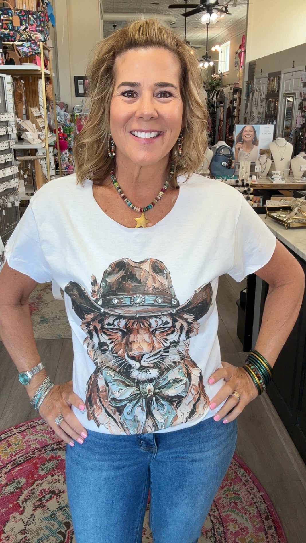 A woman smiling inside a retail shop, wearing a white graphic t-shirt with a detailed illustration of a tiger wearing a cowboy hat, paired with blue jeans.