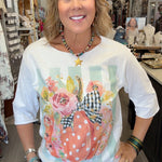 A woman smiling inside a boutique shop, wearing a white 3/4 sleeve t-shirt with a colorful graphic of a polka-dotted pumpkin, flowers, and gingham ribbon, paired with blue jeans.