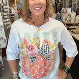 A woman smiling inside a boutique shop, wearing a white 3/4 sleeve t-shirt with a colorful graphic of a polka-dotted pumpkin, flowers, and gingham ribbon, paired with blue jeans.