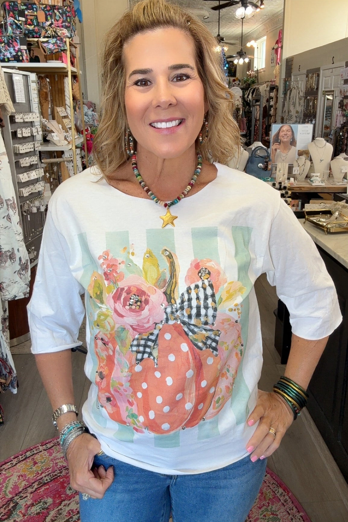 A woman smiling inside a boutique shop, wearing a white 3/4 sleeve t-shirt with a colorful graphic of a polka-dotted pumpkin, flowers, and gingham ribbon, paired with blue jeans.