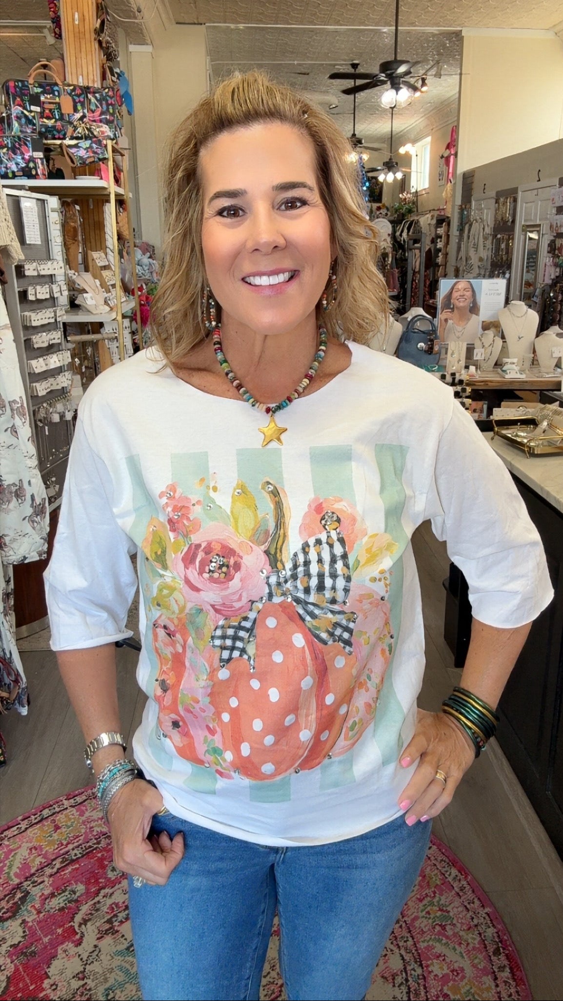 A woman smiling inside a boutique shop, wearing a white 3/4 sleeve t-shirt with a colorful graphic of a polka-dotted pumpkin, flowers, and gingham ribbon, paired with blue jeans.