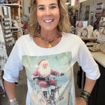 A woman wearing a white 3/4-sleeve t-shirt with a vintage-style graphic of Santa Claus riding a bicycle, standing inside a retail store.