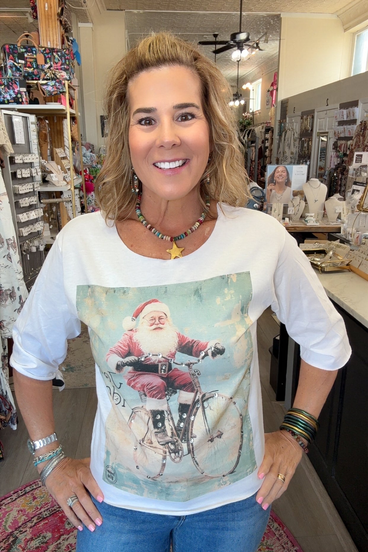 A woman wearing a white 3/4-sleeve t-shirt with a vintage-style graphic of Santa Claus riding a bicycle, standing inside a retail store.