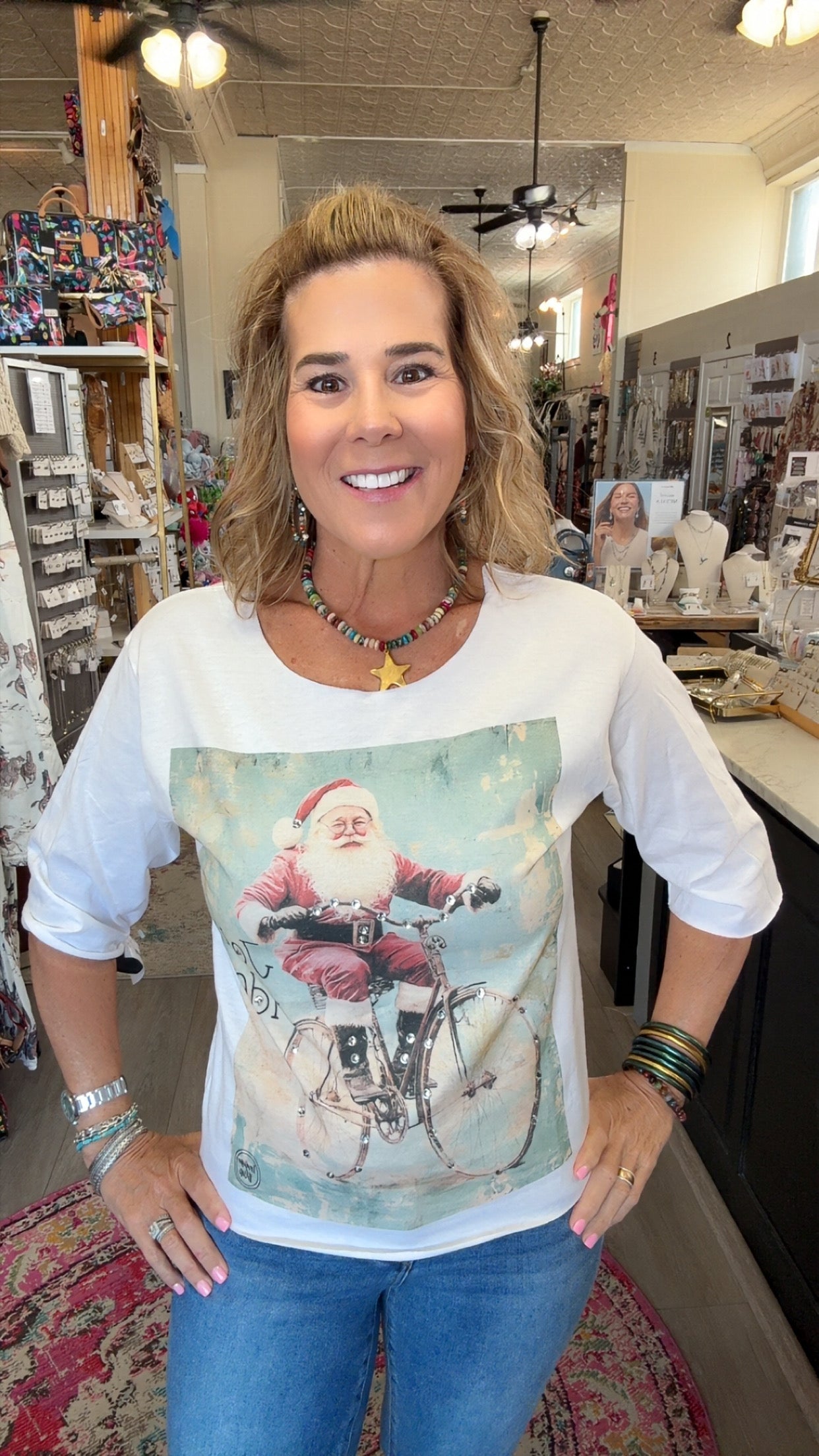A woman wearing a white 3/4-sleeve t-shirt with a vintage-style graphic of Santa Claus riding a bicycle, standing inside a retail store.