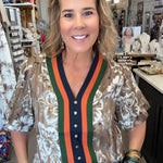 A woman posing in a boutique shop wearing a short-sleeve button-up blouse with a brown and white floral pattern, featuring a green and orange striped V-neck trim with a navy blue placket.