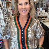 A woman posing in a boutique shop wearing a short-sleeve button-up blouse with a brown and white floral pattern, featuring a green and orange striped V-neck trim with a navy blue placket.