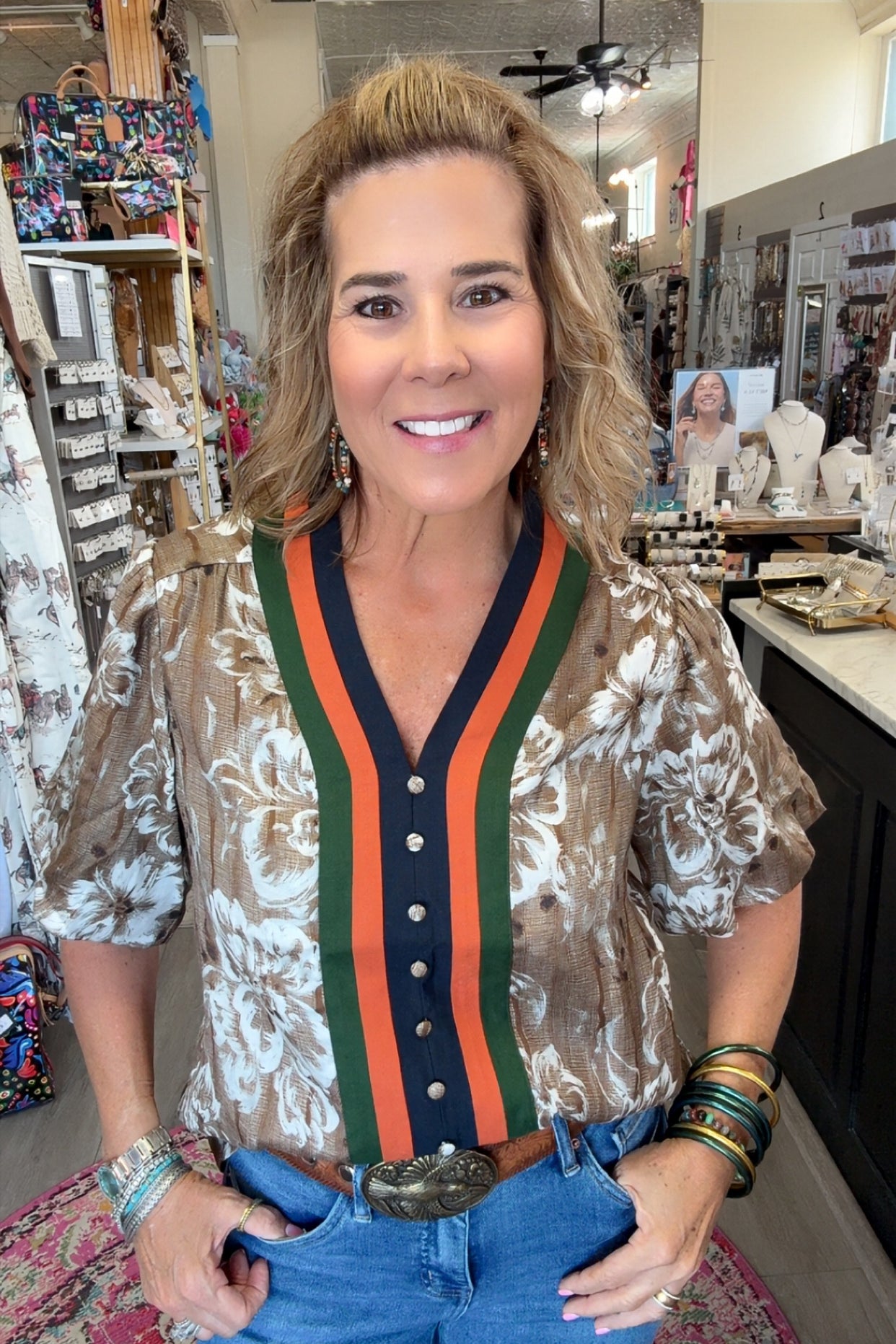 A woman posing in a boutique shop wearing a short-sleeve button-up blouse with a brown and white floral pattern, featuring a green and orange striped V-neck trim with a navy blue placket.