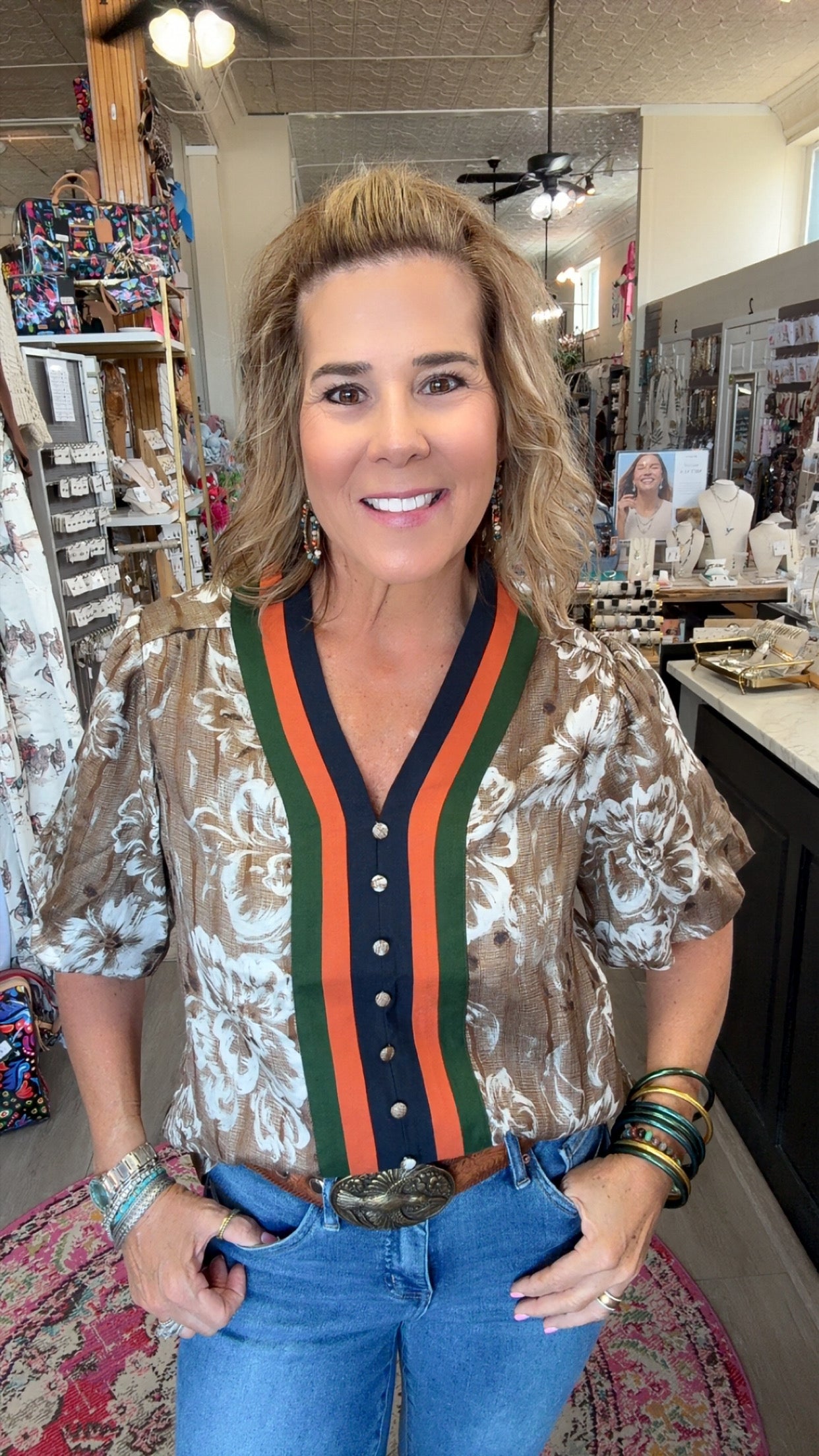 A woman posing in a boutique shop wearing a short-sleeve button-up blouse with a brown and white floral pattern, featuring a green and orange striped V-neck trim with a navy blue placket.