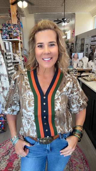 A woman posing in a boutique shop wearing a short-sleeve button-up blouse with a brown and white floral pattern, featuring a green and orange striped V-neck trim with a navy blue placket.