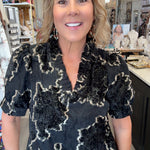 A woman with blonde hair stands in a retail store wearing a black short-sleeve blouse with a textured beige abstract floral pattern and blue jeans.