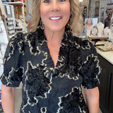 A woman with blonde hair stands in a retail store wearing a black short-sleeve blouse with a textured beige abstract floral pattern and blue jeans.
