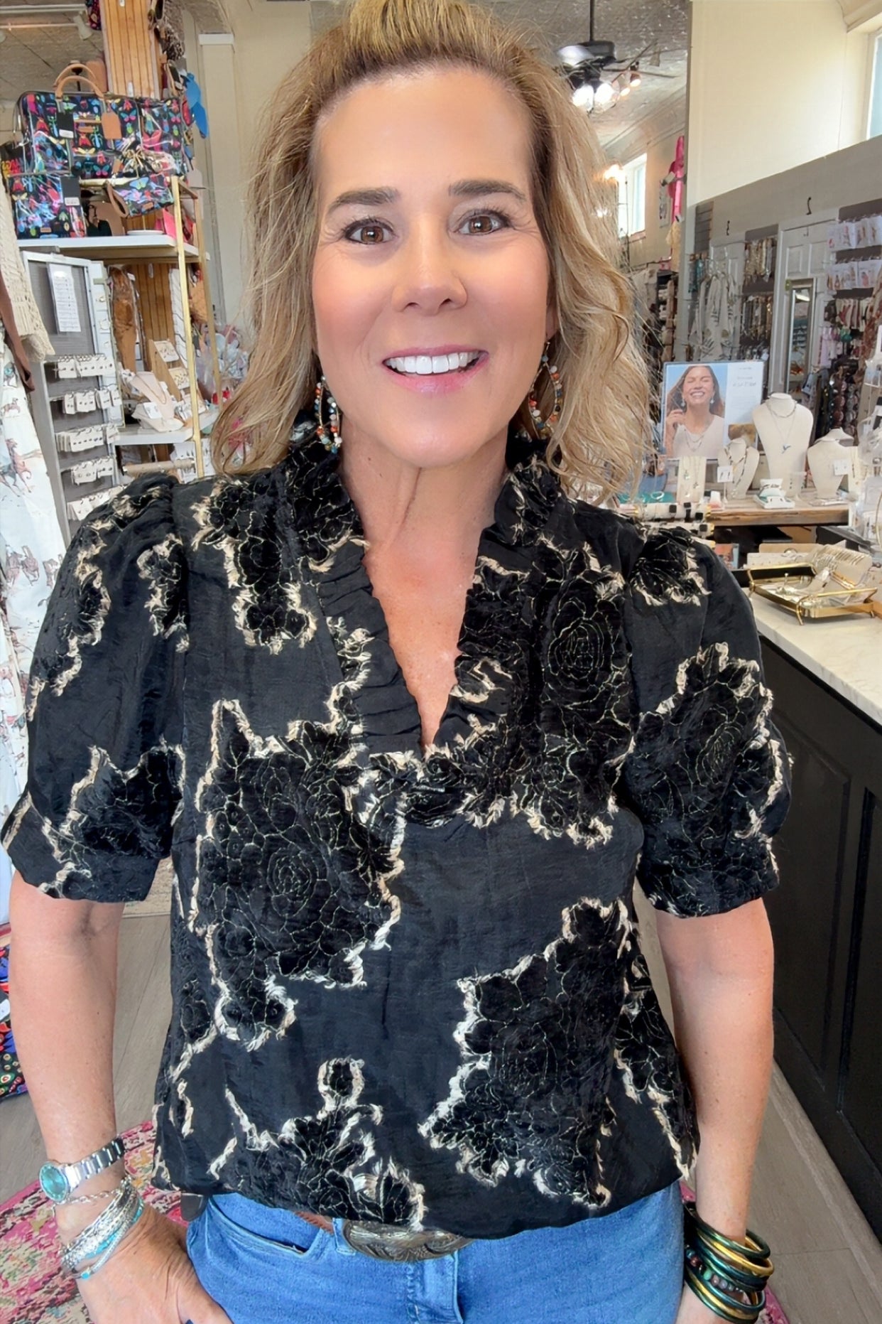 A woman with blonde hair stands in a retail store wearing a black short-sleeve blouse with a textured beige abstract floral pattern and blue jeans.