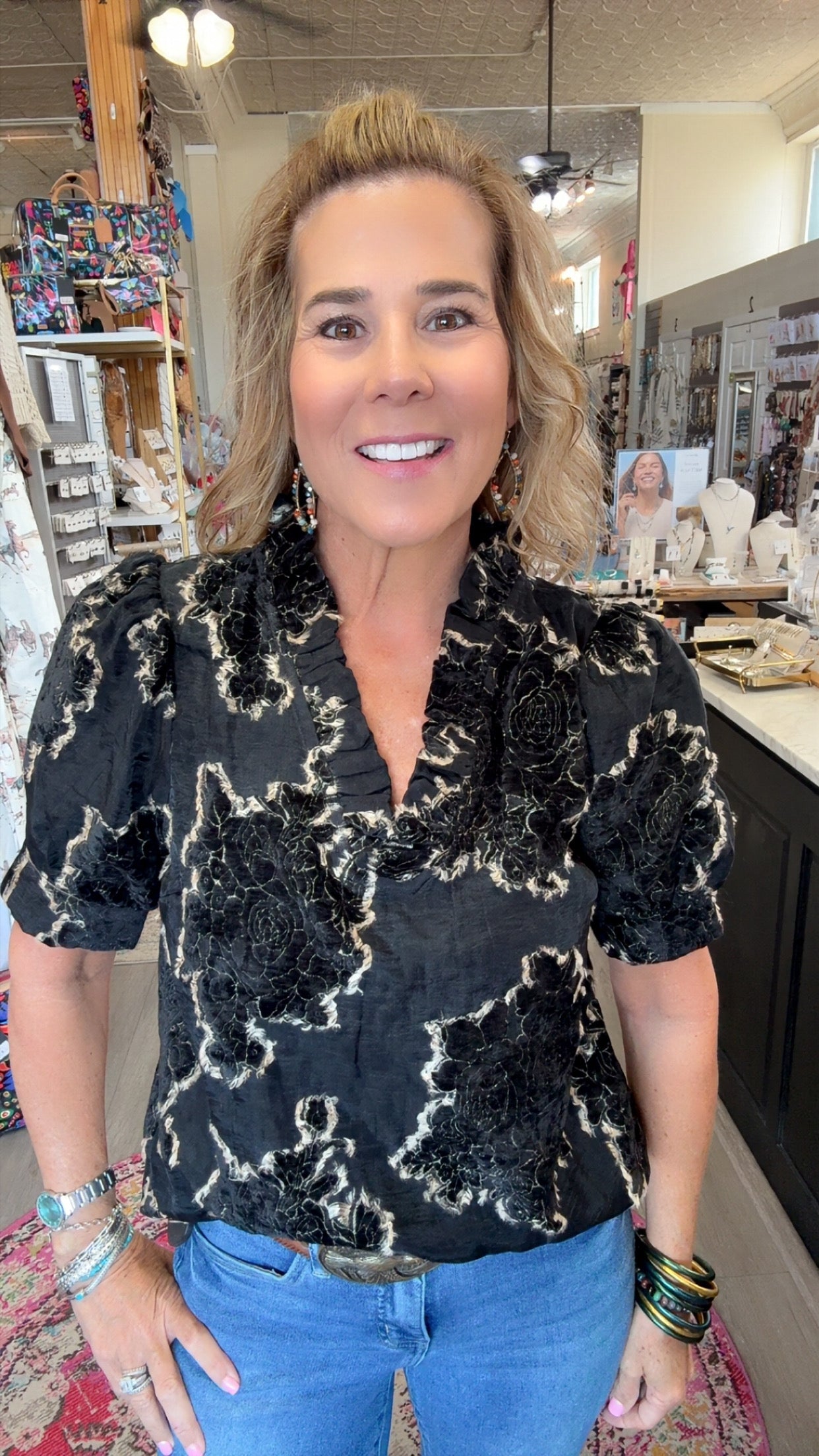 A woman with blonde hair stands in a retail store wearing a black short-sleeve blouse with a textured beige abstract floral pattern and blue jeans.