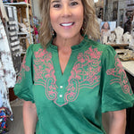 A woman wearing a green short-sleeve blouse with pink floral embroidery on the chest and sleeves, paired with blue jeans, standing inside a retail shop.