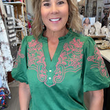 A woman wearing a green short-sleeve blouse with pink floral embroidery on the chest and sleeves, paired with blue jeans, standing inside a retail shop.
