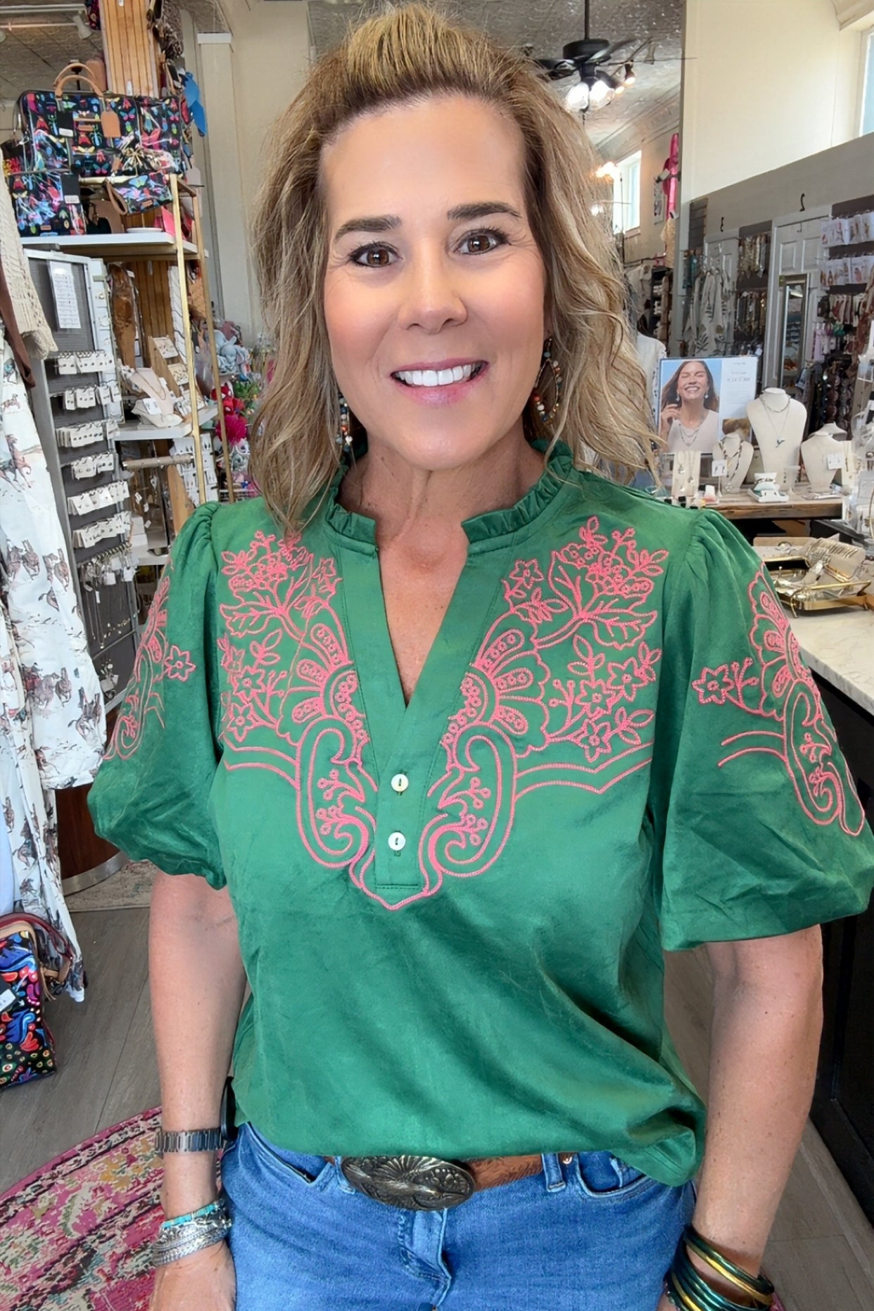 A woman wearing a green short-sleeve blouse with pink floral embroidery on the chest and sleeves, paired with blue jeans, standing inside a retail shop.