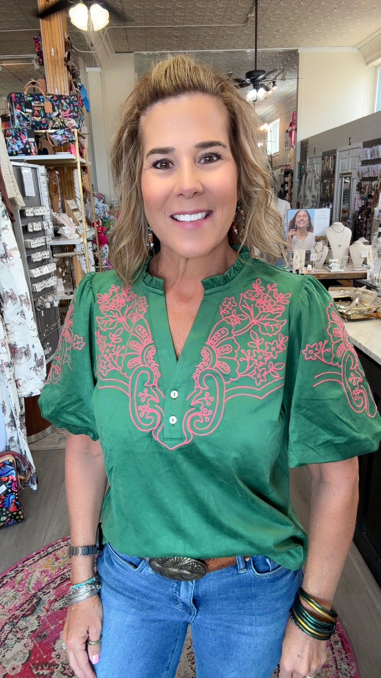 A woman wearing a green short-sleeve blouse with pink floral embroidery on the chest and sleeves, paired with blue jeans, standing inside a retail shop.