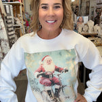 A woman wearing a white long-sleeve crewneck sweatshirt with a vintage-style graphic of Santa Claus riding a bicycle, standing in a retail shop.
