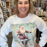 A woman wearing a white long-sleeve crewneck sweatshirt with a vintage-style graphic of Santa Claus riding a bicycle, standing in a retail shop.
