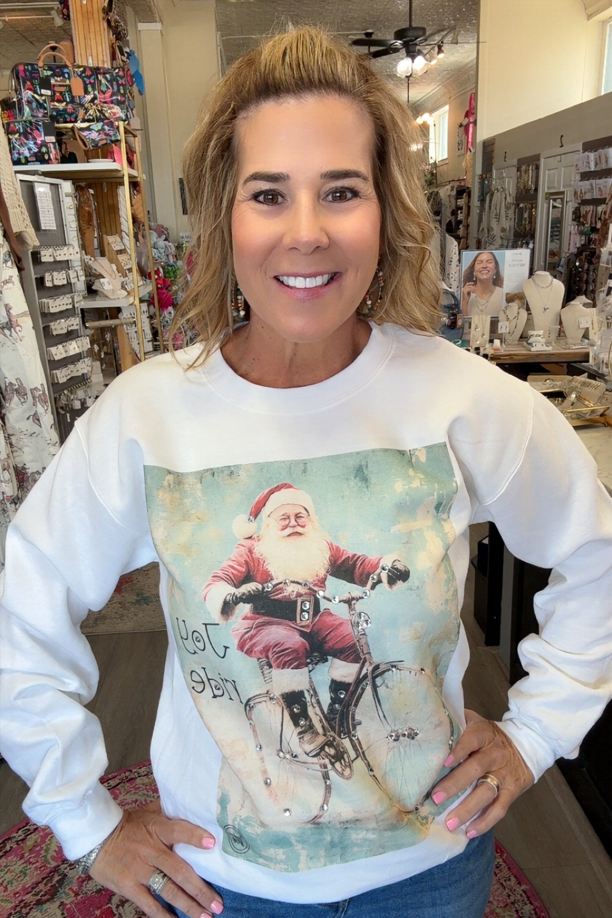 A woman wearing a white long-sleeve crewneck sweatshirt with a vintage-style graphic of Santa Claus riding a bicycle, standing in a retail shop.