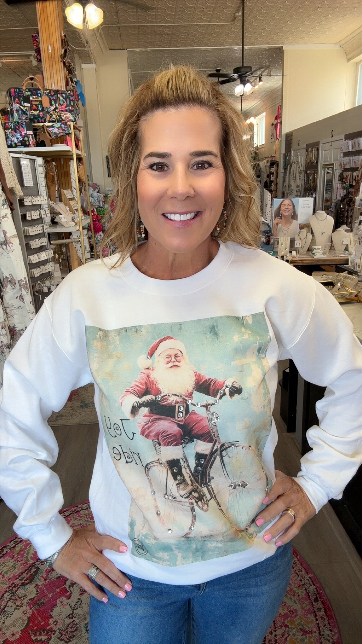 A woman wearing a white long-sleeve crewneck sweatshirt with a vintage-style graphic of Santa Claus riding a bicycle, standing in a retail shop.