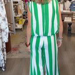 A woman viewed from behind wearing a sleeveless green and white vertically striped jumpsuit inside a retail boutique with wooden floors, clothing racks, and a patterned rug.