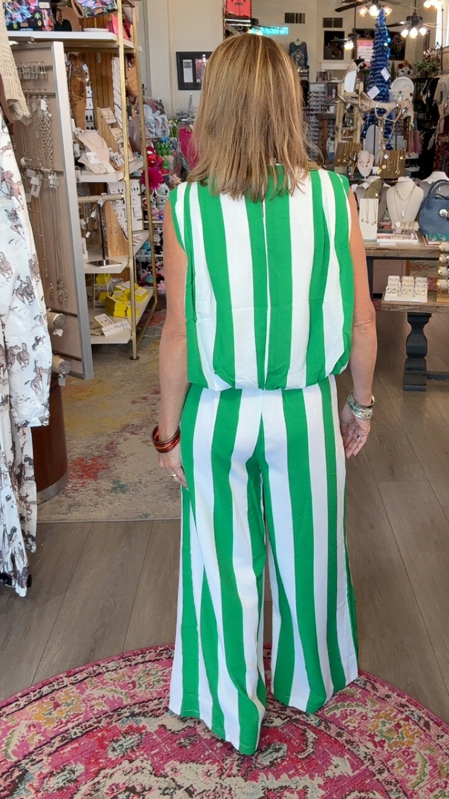 A woman viewed from behind wearing a sleeveless green and white vertically striped jumpsuit inside a retail boutique with wooden floors, clothing racks, and a patterned rug.