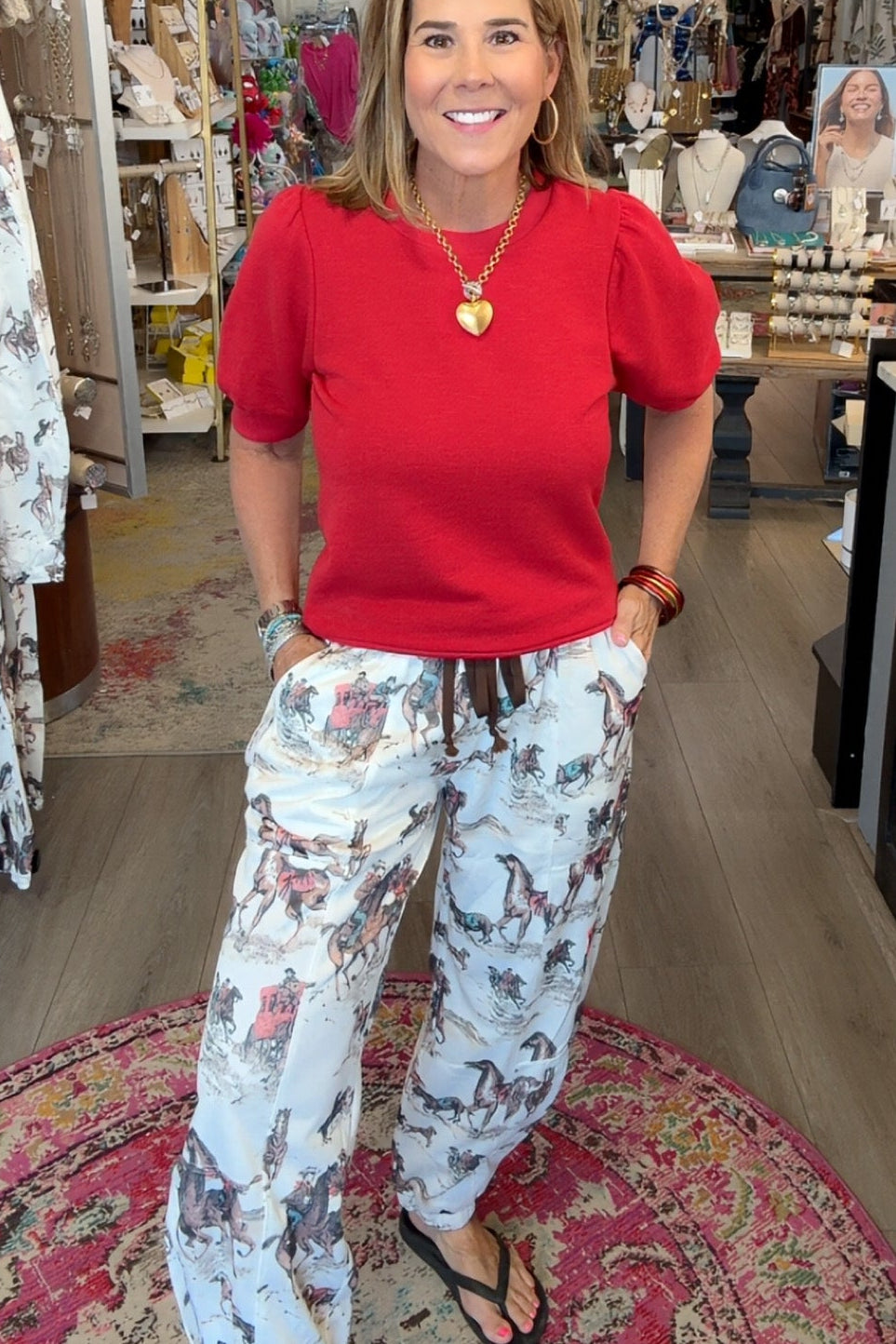 A woman stands inside a boutique shop wearing a bright red short-sleeve top and white drawstring pants with a gray and brown floral pattern. She is smiling and posing on a patterned rug, with store fixtures and clothing racks visible in the background.