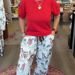 A woman stands inside a boutique shop wearing a bright red short-sleeve top and white drawstring pants with a gray and brown floral pattern. She is smiling and posing on a patterned rug, with store fixtures and clothing racks visible in the background.