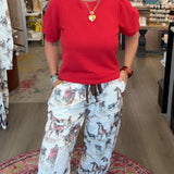 A woman stands inside a boutique shop wearing a bright red short-sleeve top and white drawstring pants with a gray and brown floral pattern. She is smiling and posing on a patterned rug, with store fixtures and clothing racks visible in the background.