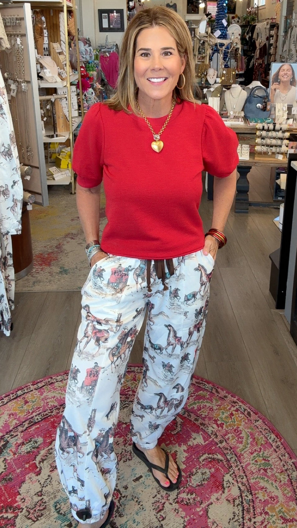 A woman stands inside a boutique shop wearing a bright red short-sleeve top and white drawstring pants with a gray and brown floral pattern. She is smiling and posing on a patterned rug, with store fixtures and clothing racks visible in the background.