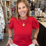 A woman smiling in a boutique shop, wearing a bright red short-sleeve puff-sleeve top and white patterned pants with a brown drawstring.