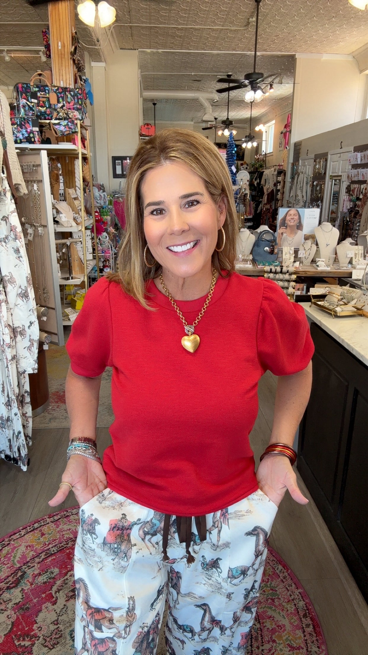 A woman smiling in a boutique shop, wearing a bright red short-sleeve puff-sleeve top and white patterned pants with a brown drawstring.
