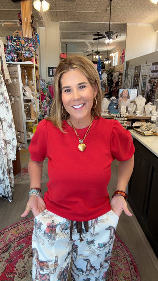 A woman smiling in a boutique shop, wearing a bright red short-sleeve puff-sleeve top and white patterned pants with a brown drawstring.