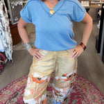 A woman posing in a boutique shop, wearing a light blue short-sleeve top and beige wide-leg pants with a colorful flamingo and floral print.