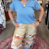 A woman posing in a boutique shop, wearing a light blue short-sleeve top and beige wide-leg pants with a colorful flamingo and floral print.