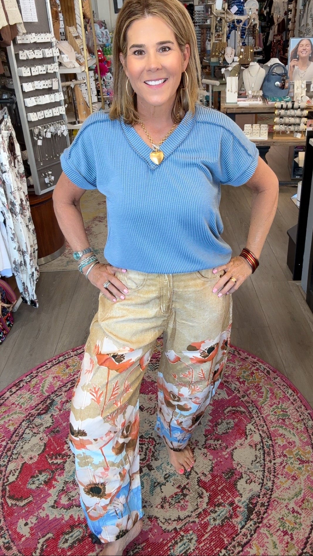A woman posing in a boutique shop, wearing a light blue short-sleeve top and beige wide-leg pants with a colorful flamingo and floral print.