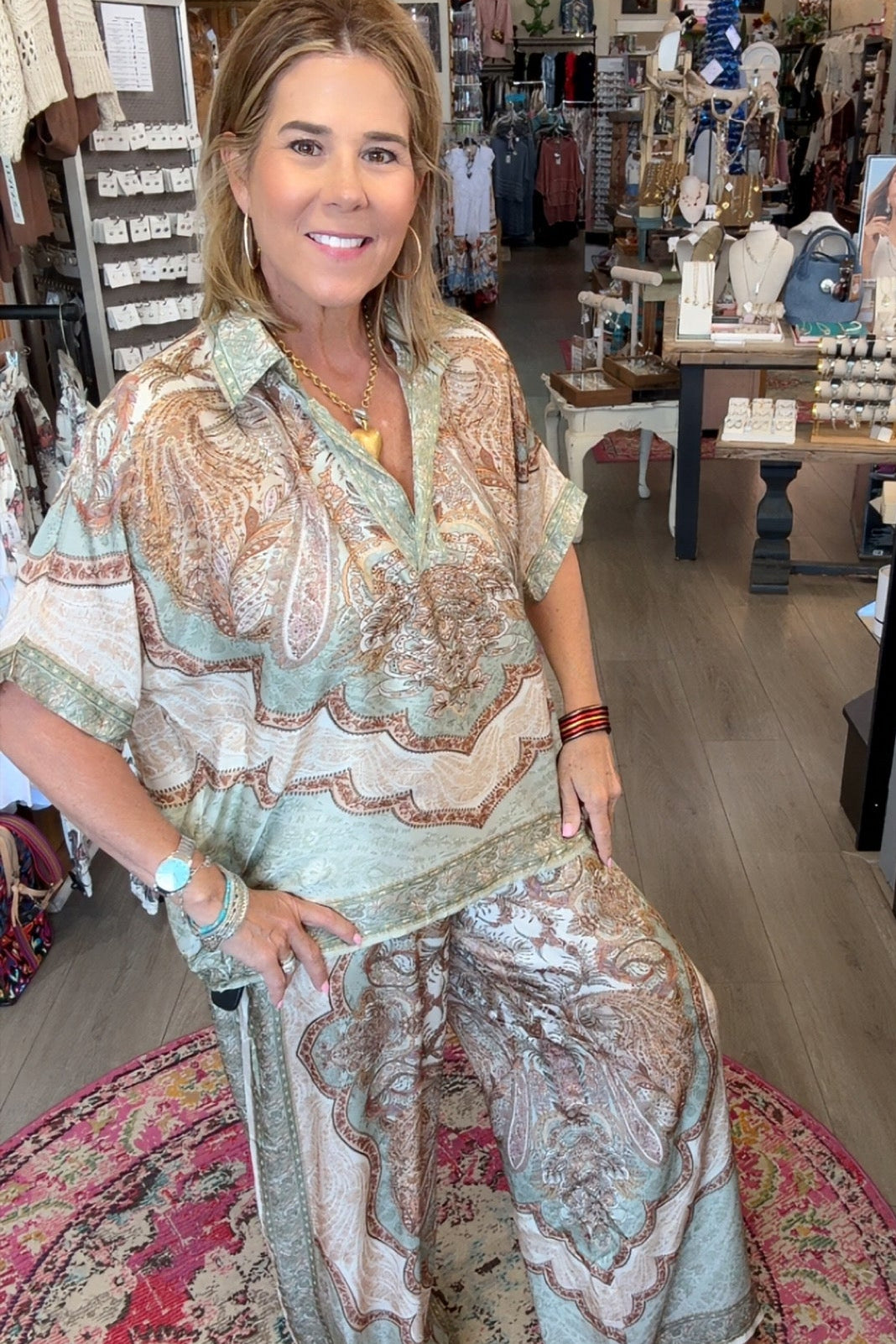 A woman posing in a bohemian-style clothing boutique, wearing a light sage green and beige paisley print two-piece set consisting of a short-sleeve button-up shirt and matching wide-leg pants.