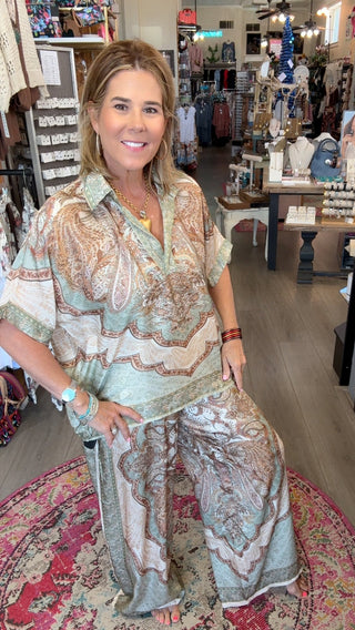 A woman posing in a bohemian-style clothing boutique, wearing a light sage green and beige paisley print two-piece set consisting of a short-sleeve button-up shirt and matching wide-leg pants.